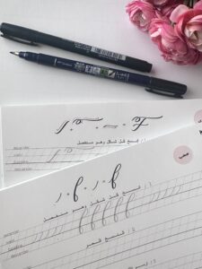 Close-up of two Tombow brush pens placed beside two sheets of modern English calligraphy practice worksheets, featuring elegant letter forms and detailed guidelines for beginners and advanced learners. The scene is complemented by a cluster of pink flowers, adding a soft and creative ambiance