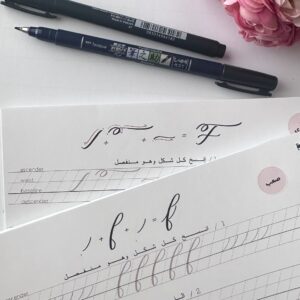 Close-up of two Tombow brush pens placed beside two sheets of modern English calligraphy practice worksheets, featuring elegant letter forms and detailed guidelines for beginners and advanced learners. The scene is complemented by a cluster of pink flowers, adding a soft and creative ambiance