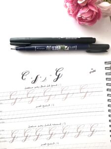  Modern calligraphy practice worksheet focused on the 'Descending Loop' technique, with a Tombow brush pen placed on the sheet. The worksheet, written in both Arabic and English, includes practice lines for mastering the descending loop strokes. A pink floral decoration is visible in the background, adding a creative and elegant touch