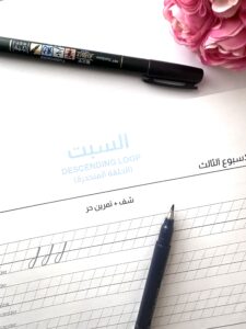  “Modern calligraphy practice worksheet focused on the 'Descending Loop' technique, with a Tombow brush pen placed on the sheet. The worksheet, written in both Arabic and English, includes practice lines for mastering the descending loop strokes. A pink floral decoration is visible in the background, adding a creative and elegant touch“