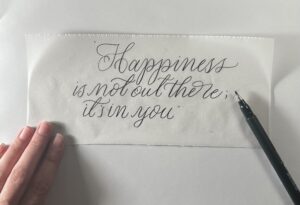 The Alt Text for this image could be: "Handwritten calligraphy on a piece of paper reading, 'Happiness is not out there; it's in you,' created with a Fudenosuke brush pen (hard tip) placed on the right side. A hand is holding the paper in the lower left corner." 