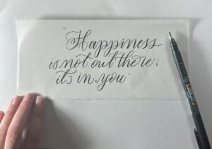 The Alt Text for this image could be: "Calligraphy quote written in black ink on a piece of translucent paper, reading 'Happiness is not out there; it's in you.' A hand is holding the paper in the lower left corner, and a Fudenosuke brush pen (soft tip) is placed on the right side of the paper." 